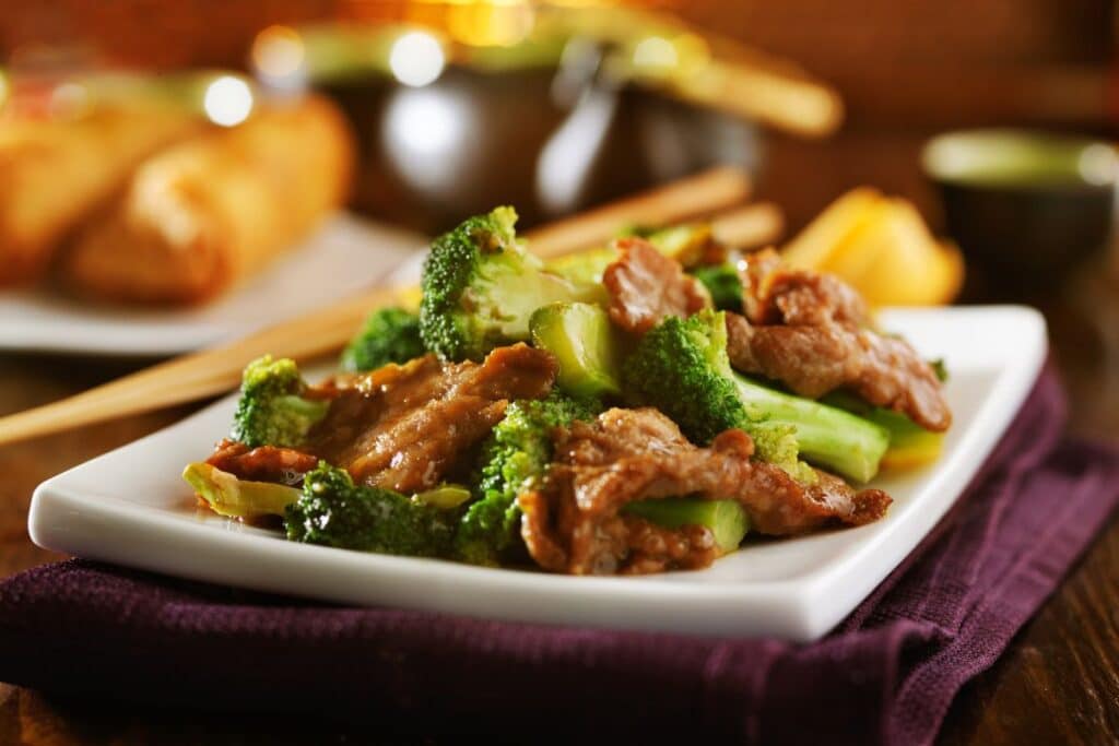 A rectangular white plate with beef and broccoli stir-fry, a classic homemade Chinese food, sits on a dark serviette. In the blurred background, spring rolls, chopsticks, and a cup are visible.