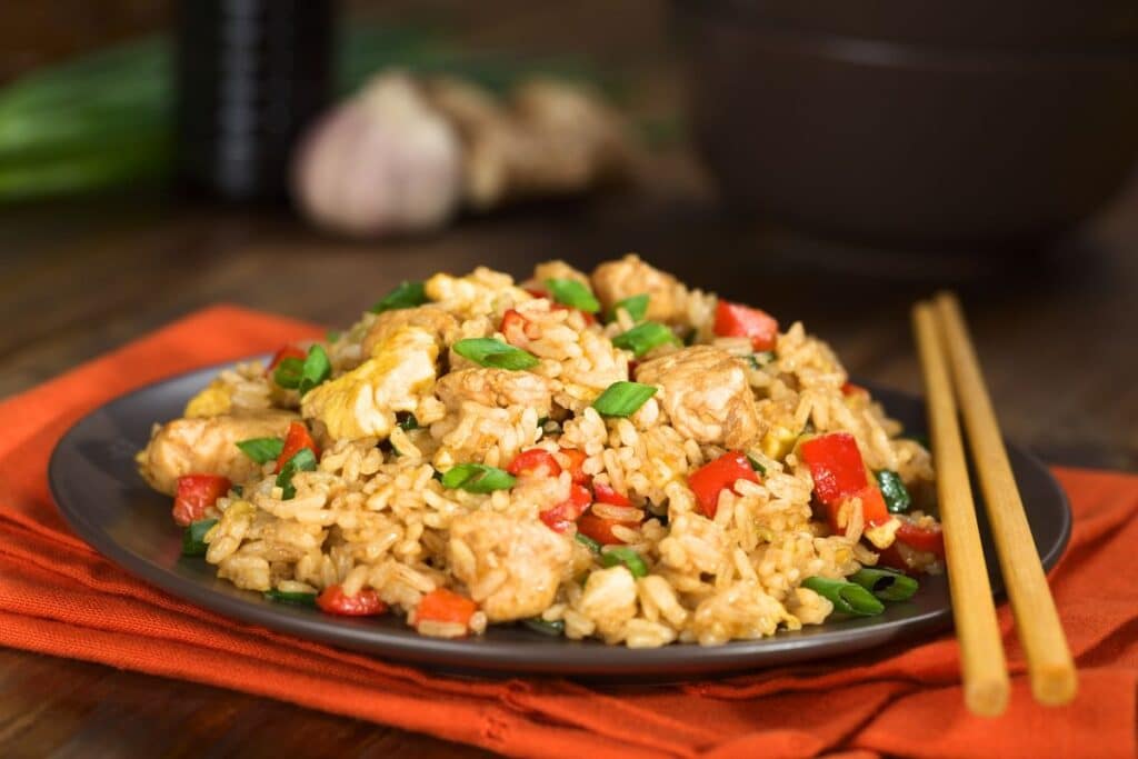 A plate of homemade Chinese food featuring chicken fried rice with diced red peppers, scrambled eggs, and spring onions, served with chopsticks on an orange serviette, and blurred vegetables in the background.