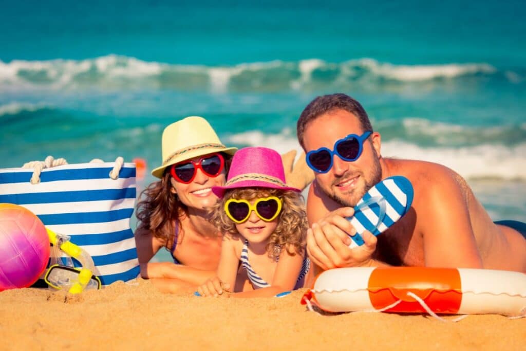 A smiling family of three wearing colourful hats and heart-shaped sunglasses lies on the sand at the beach with the sea behind them, enjoying a day of fun while keeping beach safety in mind. A striped beach bag and beach toys are nearby.