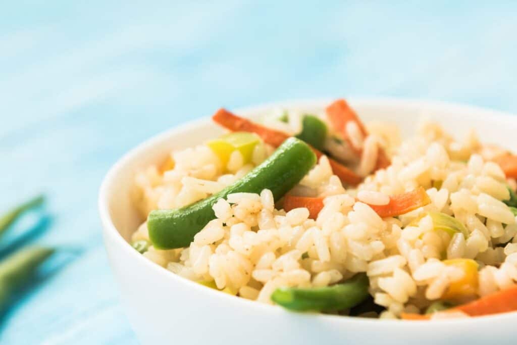 A close-up of a white bowl filled with cooked rice mixed with green beans, carrots, and other vegetables, capturing the essence of home-made Chinese food against a light blue background.