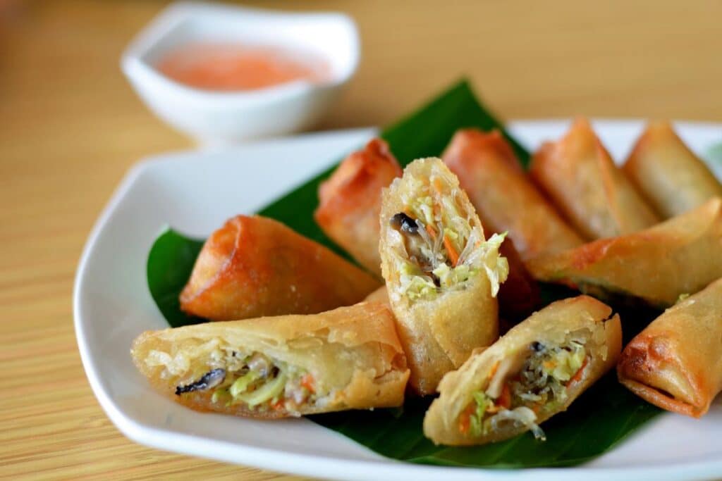 A plate of golden-brown vegetable spring rolls, some cut open to show the filling, is arranged on a green leaf—a delightful example of homemade Chinese food. A small dish of dipping sauce sits in the background.