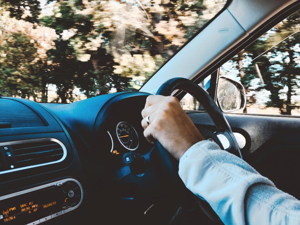 A person’s left hand gripping a steering wheel inside a car, with dashboard details and blurred green trees outside the window, capturing the feel of a long car journey. The photo is taken from the front passenger seat.