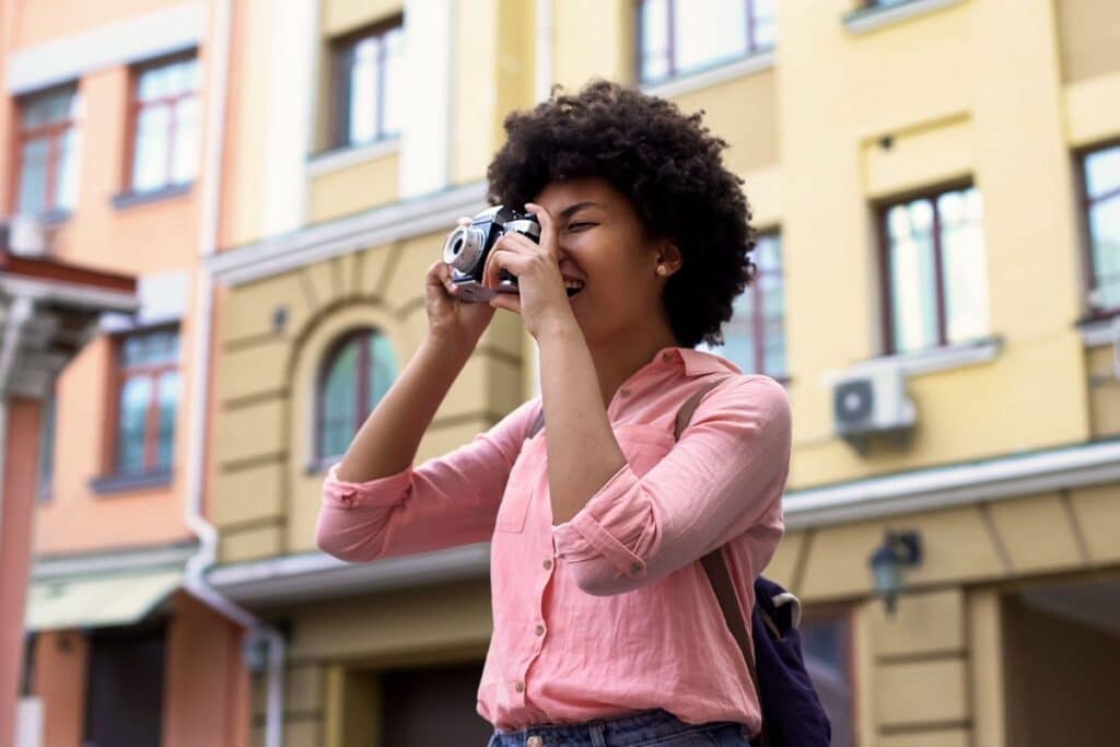 A young woman with curly hair, wearing a light pink shirt, smiles as she captures memories during her gap year, taking a photo with a camera outside in an urban area with colourful buildings in the background.
