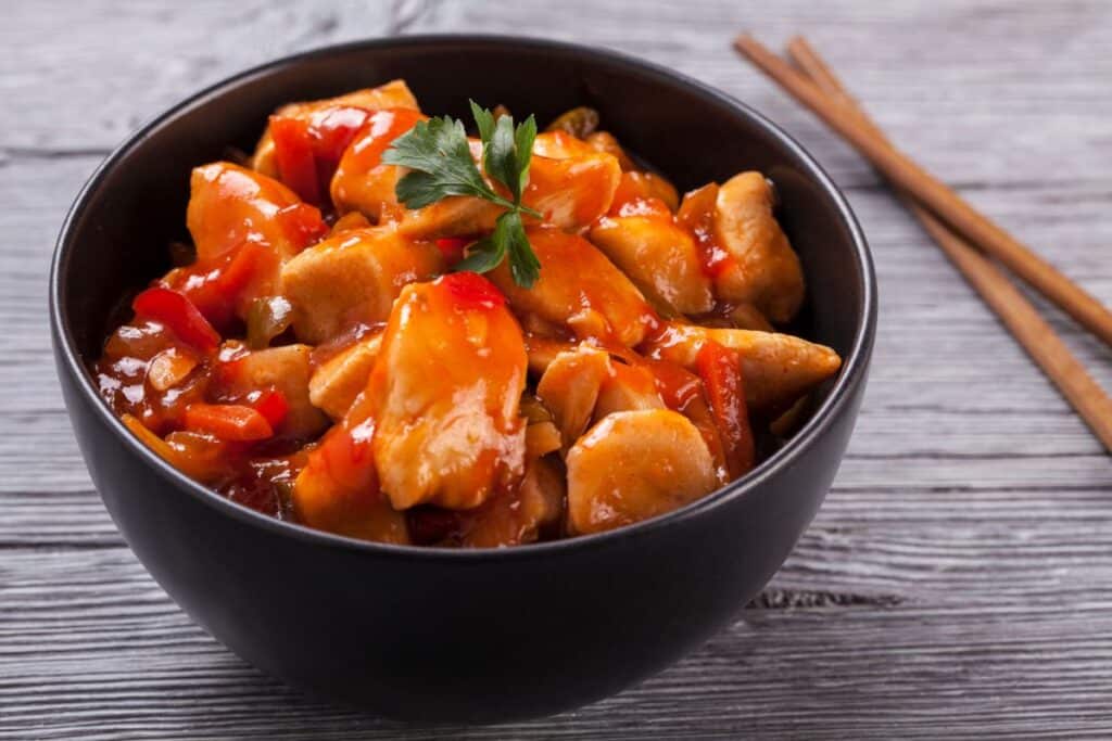 A black bowl filled with homemade Chinese food—sweet and sour chicken—garnished with a sprig of parsley, sits on a wooden table with a pair of chopsticks beside it.