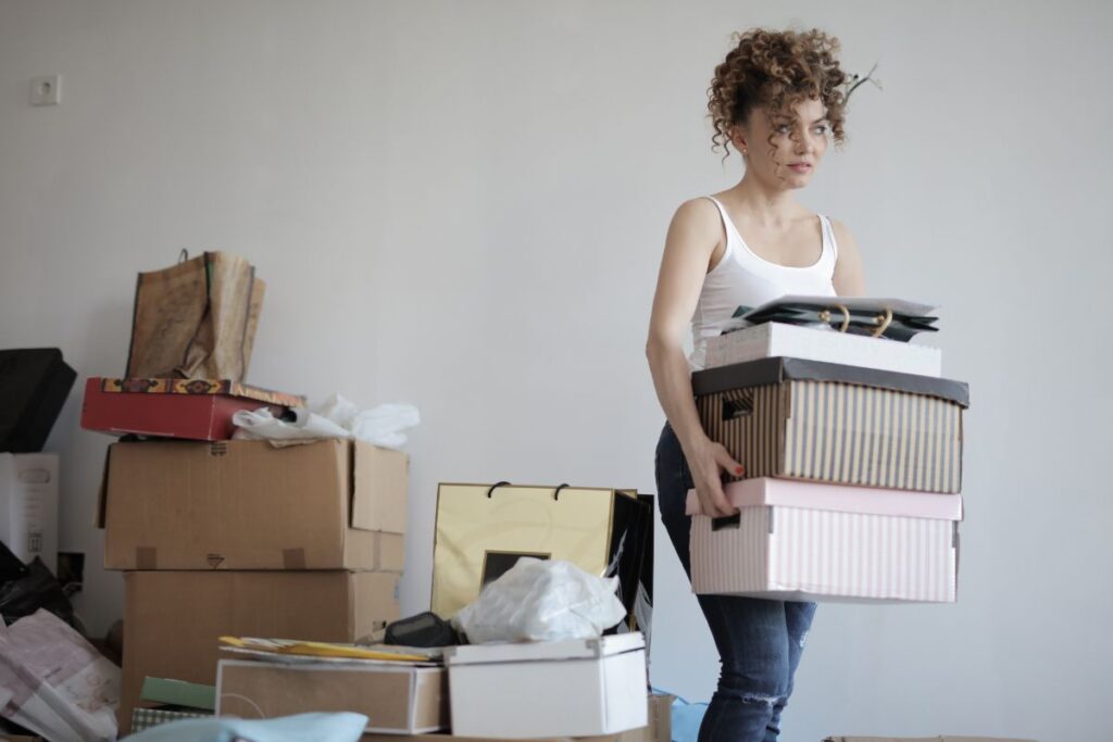 A woman with curly hair in a white vest holds a stack of boxes while standing in a room cluttered with various moving boxes and bags, preparing items for storage.