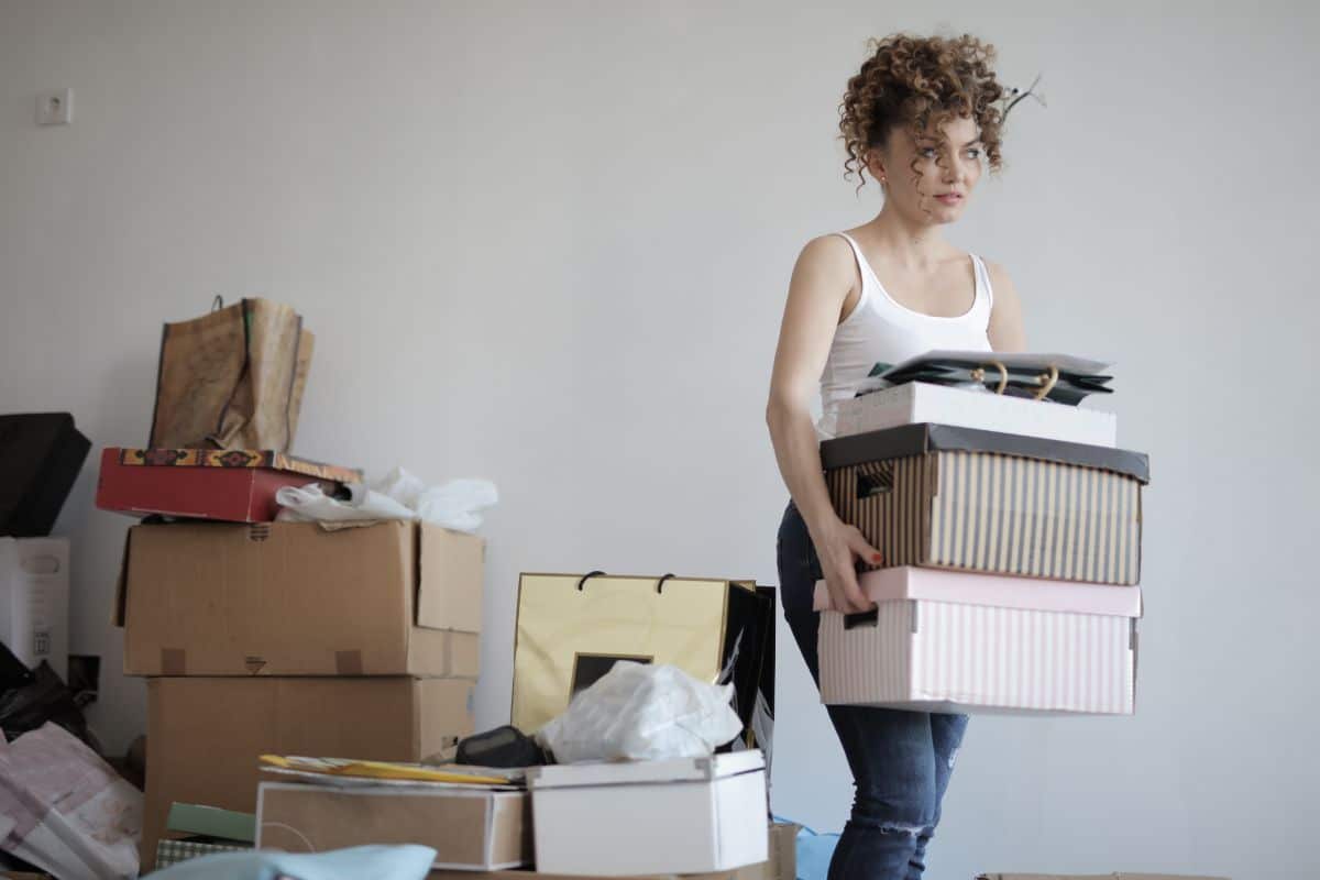 A woman with curly hair in a white vest holds a stack of boxes while standing in a room cluttered with various moving boxes and bags, preparing items for storage.