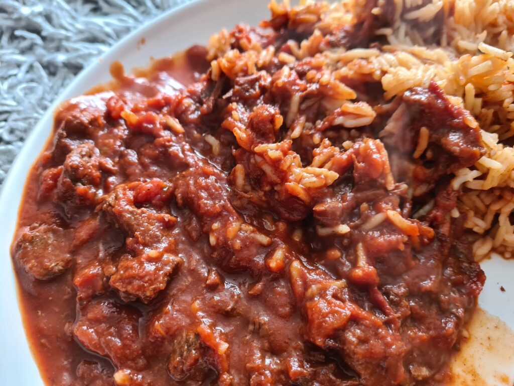 A close-up of a plate with slow cooker beef curry in a rich, red tomato sauce next to a serving of cooked rice. The dish appears hearty and flavourful, with tender chunks of meat and visible grains of rice.