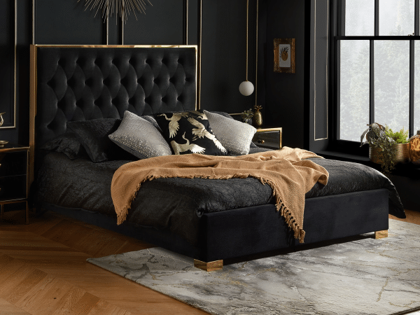 A stylish bedroom with a dark tufted bed frame, gold accents, a tan blanket, bird-patterned pillows, dark walls, large windows, bedside tables, hanging lights, and a light patterned rug on wooden flooring.