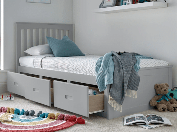 A grey bed frame with drawers underneath, two open and one closed, sits against a white wall. The bed has white and blue bedding with a throw blanket and cushions. Nearby are a stuffed bear, books, and a colourful rug on the floor.