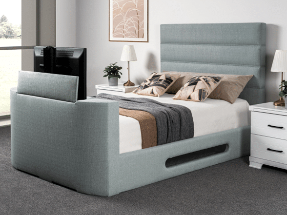 A modern bedroom with a light grey upholstered bed frame featuring a tall headboard and footboard, a TV at the foot of the bed, two white bedside tables with lamps, and neutral-toned bedding accented by decorative cushions.