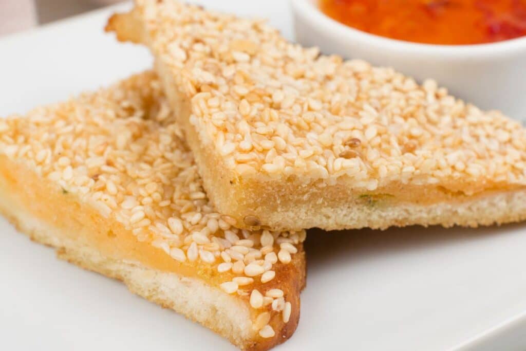 Two triangular slices of homemade prawn toast on a white plate, topped with golden sesame seeds, with a small bowl of sweet chilli dipping sauce in the background.
