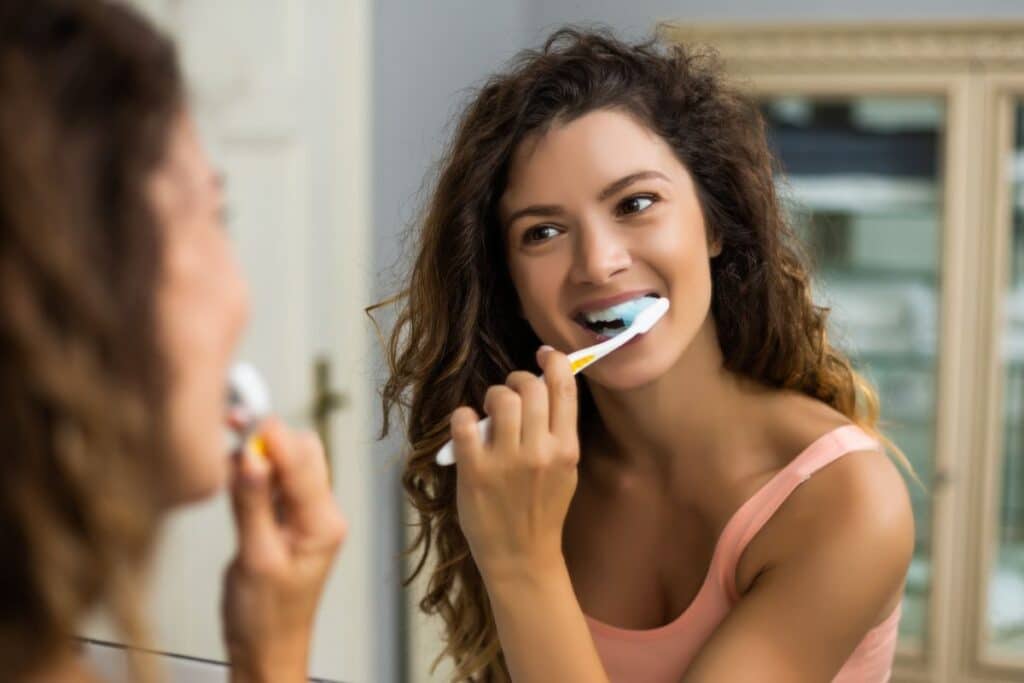 A woman with curly hair smiles whilst brushing her teeth, looking at herself in the bathroom mirror. She is wearing a pink vest top. Her bright teeth add to her cheerful morning routine.