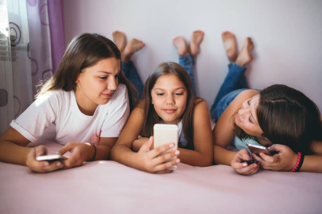 Three girls lying on a bed, legs up, holding and looking at mobiles together, smiling and relaxed in a brightly lit room as they scroll through social media.