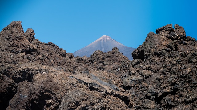A distant volcanic peak with a snow-capped top rises above Tenerife's dark, jagged volcanic rocks under a clear blue sky.