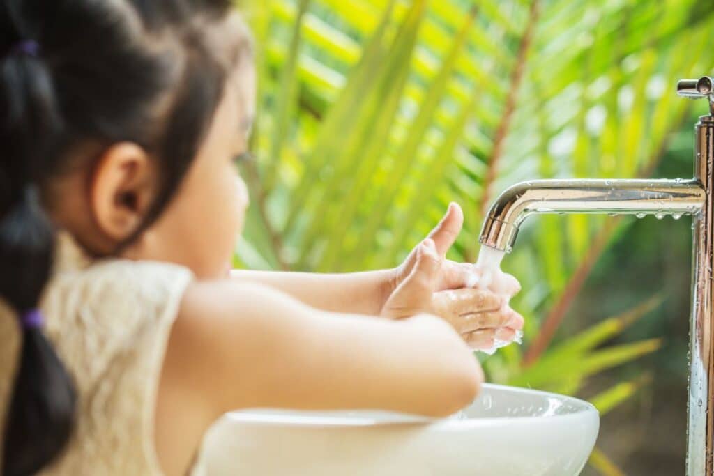 A young child practises personal hygiene by washing her hands under a running tap in a white sink, with green palm leaves visible in the background.