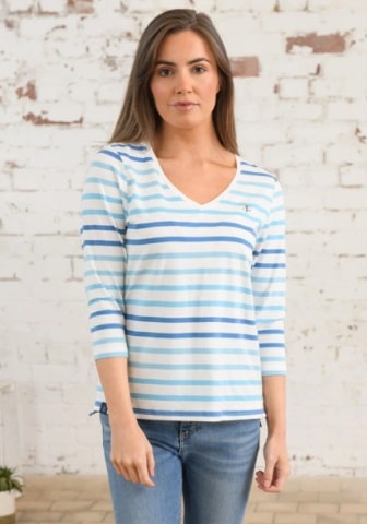 A woman with long brown hair wears a light blue and white striped, long-sleeved shirt and blue jeans, standing in front of a white brick wall—the perfect outfit for walking your dog.