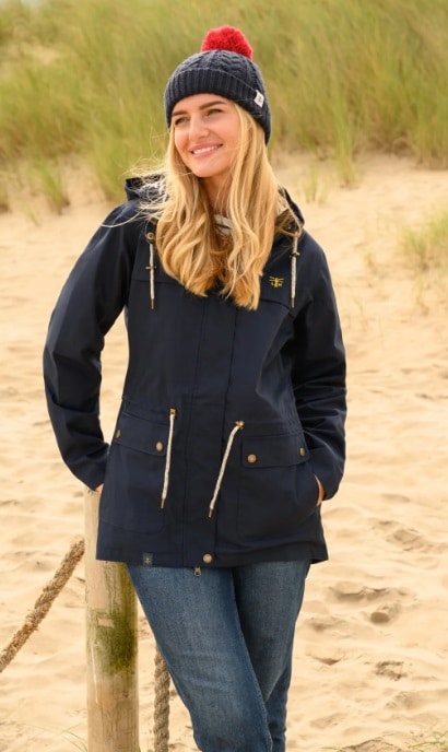 A woman wearing a navy jacket, blue jeans, and a navy knitted hat with a red bobble stands outdoors on a sandy path with tall grass—smiling and leaning against a wooden post in the perfect outfit for walking your dog.