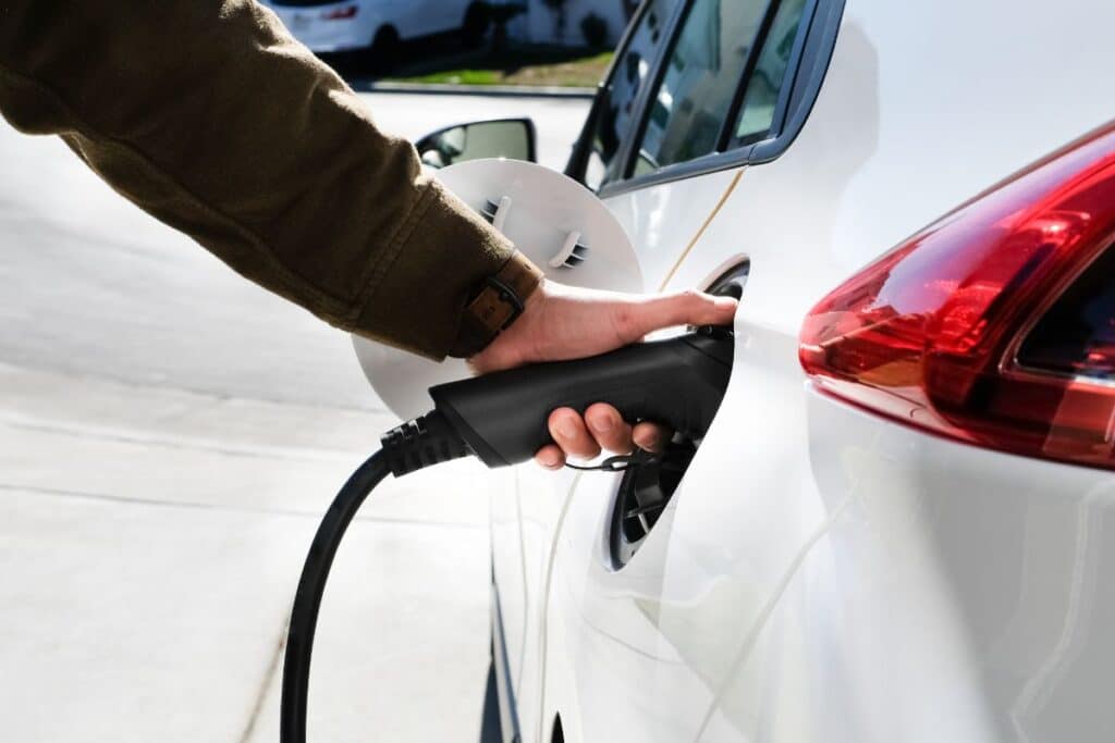 A person’s hand plugs an EV charger cable into a white electric car parked outdoors. The charging port’s cover is open, and the vehicle’s rear light is visible.