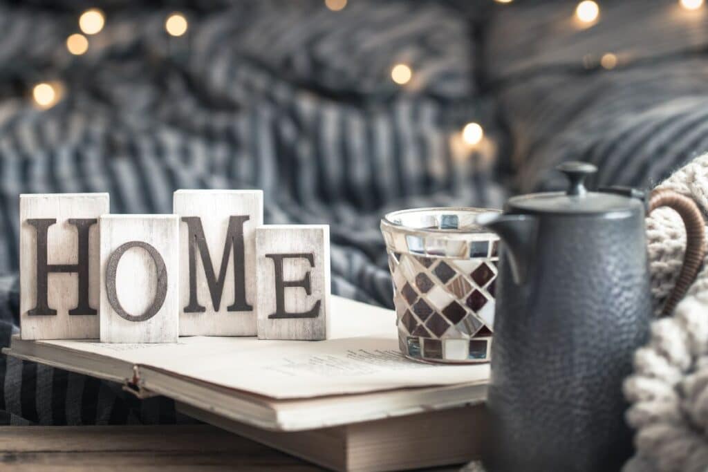 Wooden blocks spelling “HOME” sit on a book beside a patterned candle holder and dark teapot, with cosy blankets and fairy lights in the blurred background—perfect inspiration to spruce up your home.