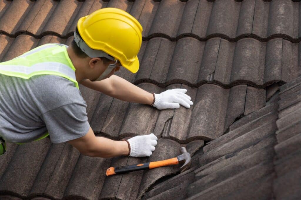 A construction worker wearing a yellow hard hat, high-visibility vest, and white gloves repairs roof tiles with a hammer on the brown tiled roof of a house.