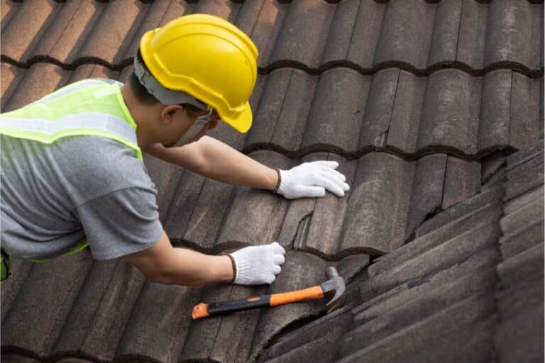 A construction worker wearing a yellow hard hat, high-visibility vest, and white gloves repairs roof tiles with a hammer on the brown tiled roof of a house.