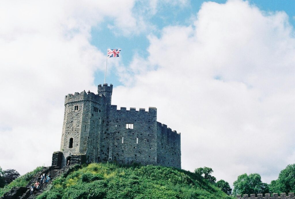 An historic stone castle with battlements stands on a grassy hill under a partly cloudy UK sky. A large Union Flag flies from the top of the castle, whilst a few people are visible near the base of the structure.