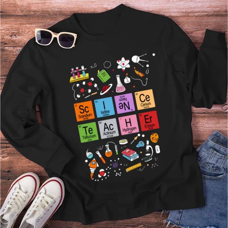 A black jumper with colourful science-themed graphics, including laboratory equipment, molecules, and six periodic table elements spelling “SCIENCER”—perfect for the classroom. White trainers and sunglasses are beside the jumper on a wooden surface.