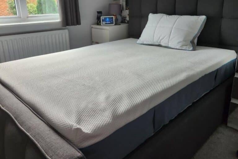 A neatly made double bed with a white textured mattress cover, ideal for a light sleeper, two pillows, and a grey upholstered headboard, positioned next to a window with grey curtains and a bedside table holding a clock and other items.