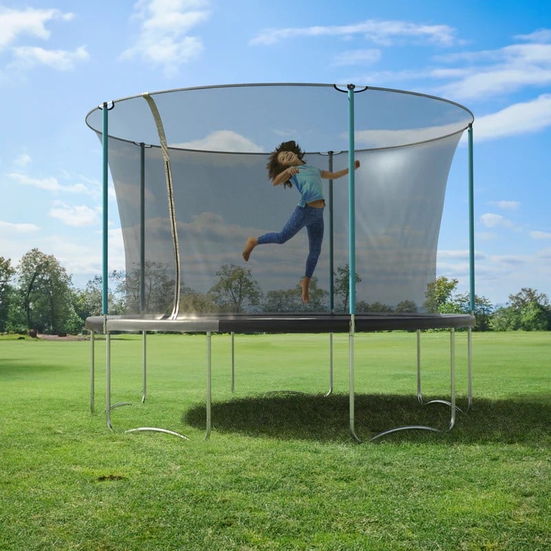 A child with long hair, wearing a blue shirt and jeans, jumps high on a safety net trampoline. Trampolines like this one are set up on a grassy field under a bright blue sky with scattered clouds.