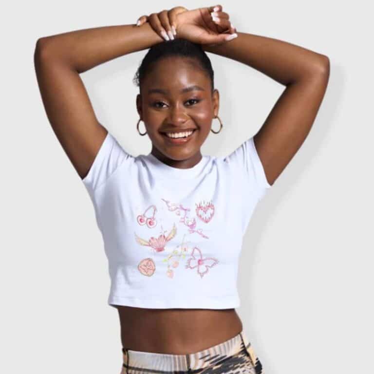 A smiling woman with arms raised above her head wears a white crop top with colourful abstract designs and patterned leggings, channelling Y2K fashion. She has hoop earrings and a neat bun hairstyle, standing against a plain light background.