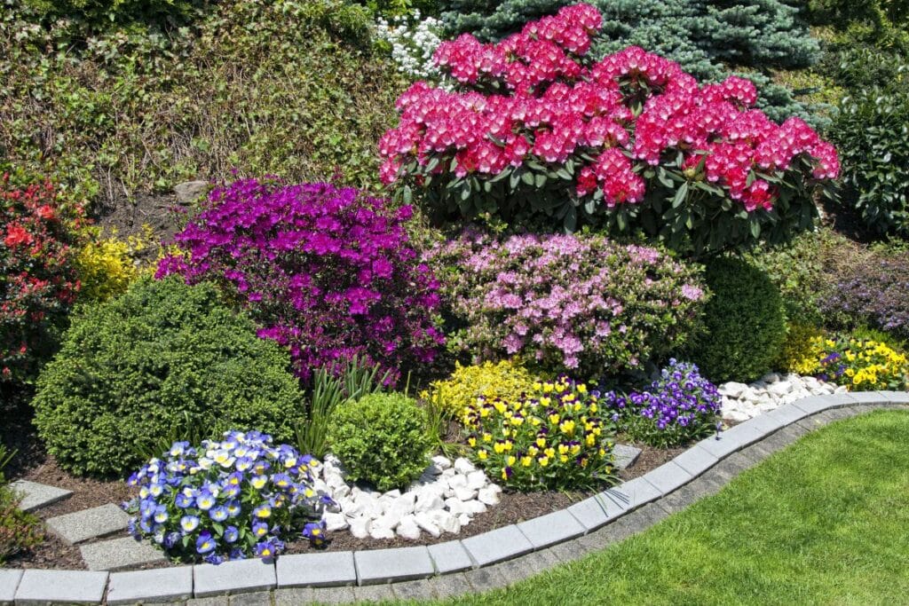 A vibrant garden bordered by grey bricks, featuring blooming pink, purple, yellow, and blue flowers, lush green shrubs, and a neatly trimmed garden lawn in the foreground.