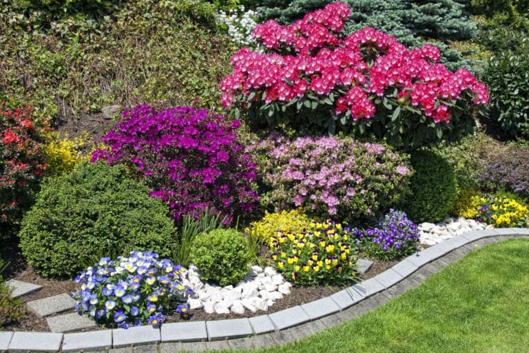 A vibrant garden bordered by grey bricks, featuring blooming pink, purple, yellow, and blue flowers, lush green shrubs, and a neatly trimmed garden lawn in the foreground.