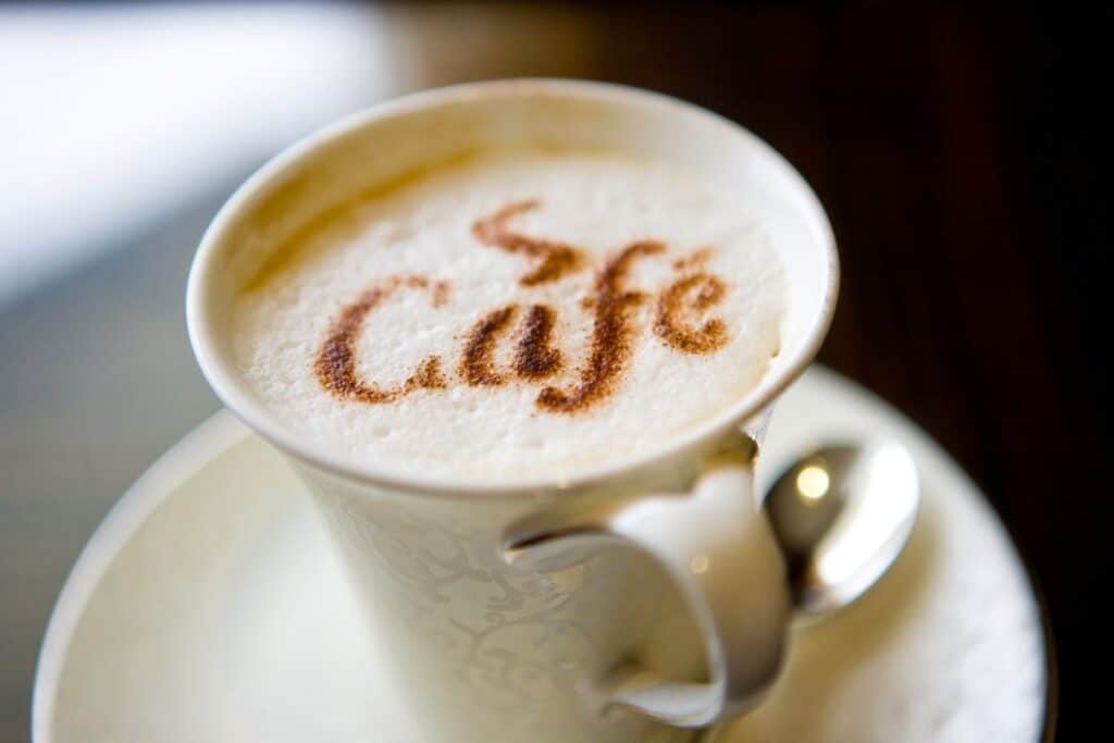 A cup of cappuccino with the word "Café" written in cocoa powder on the frothy milk sits on a white saucer with a spoon, atop practical furniture that complements any cosy coffee nook.