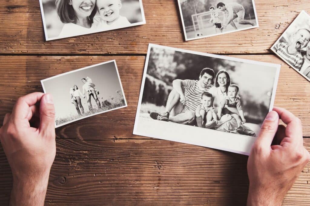 Hands holding black-and-white family photos over a rustic wooden table, with additional scattered photos showing smiling people and children—capturing cherished memories across generations.