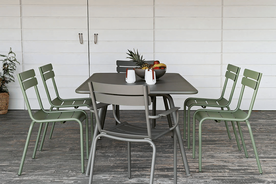 A practical furniture choice, this patio dining set features a rectangular table and six green chairs on a wooden decking. The table displays a bowl of fruit, a pineapple, and salt and pepper shakers. A plant accents the white wall in the background.