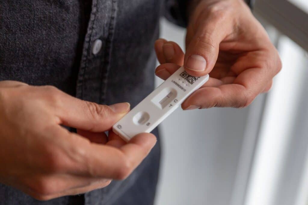 A person holds a lateral flow test cassette for COVID-19, showing a single line in the control area that indicates a negative result. The individual is wearing a dark shirt.