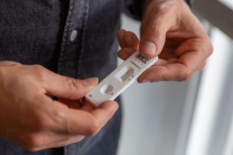 A person holds a lateral flow test cassette for COVID-19, showing a single line in the control area that indicates a negative result. The individual is wearing a dark shirt.