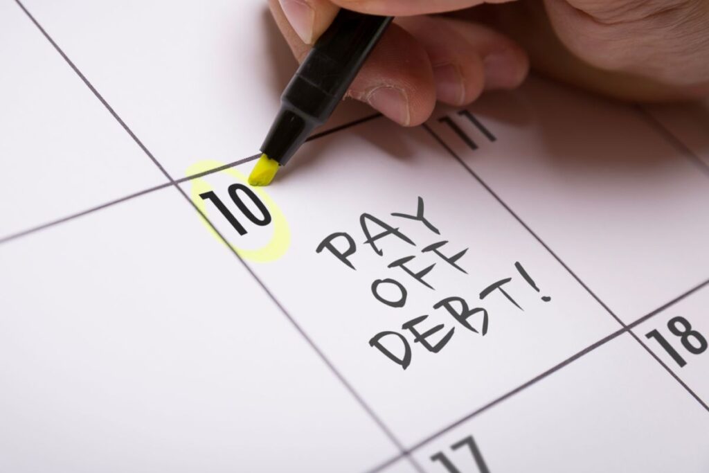 A hand circles the 10th day on a calendar with a yellow marker, highlighting the word “debt” as “PAY OFF DEBT!” is written on that date.
