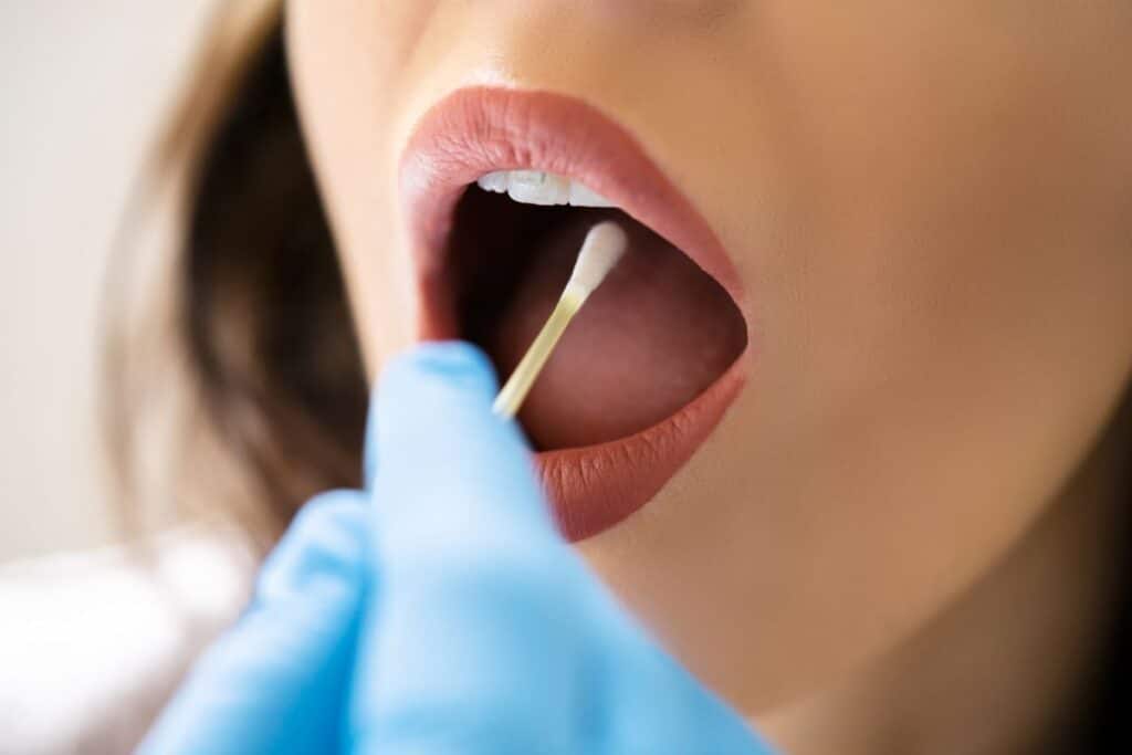 A close-up of a person’s mouth open wide as a gloved hand uses a cotton bud to collect a sample for saliva-based tests from inside their cheek.
