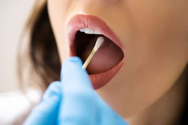 A close-up of a person’s mouth open wide as a gloved hand uses a cotton bud to collect a sample for saliva-based tests from inside their cheek.