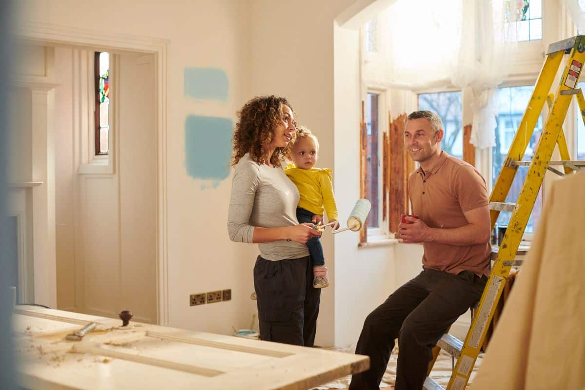A woman holding a toddler and a paint roller stands next to a man sitting on a ladder in a sunlit room, surrounded by renovation materials—a lively scene of updating your home with fresh paint samples on the wall.
