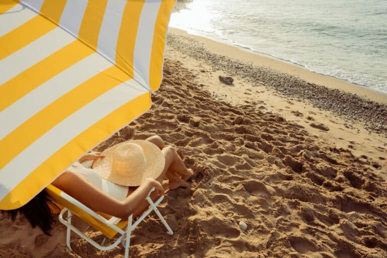 A person enjoys a beach holiday, relaxing on a sandy shore under a yellow and white striped parasol, sitting on a chair with a wide-brimmed hat and gazing at the sparkling sea in the sunlight.