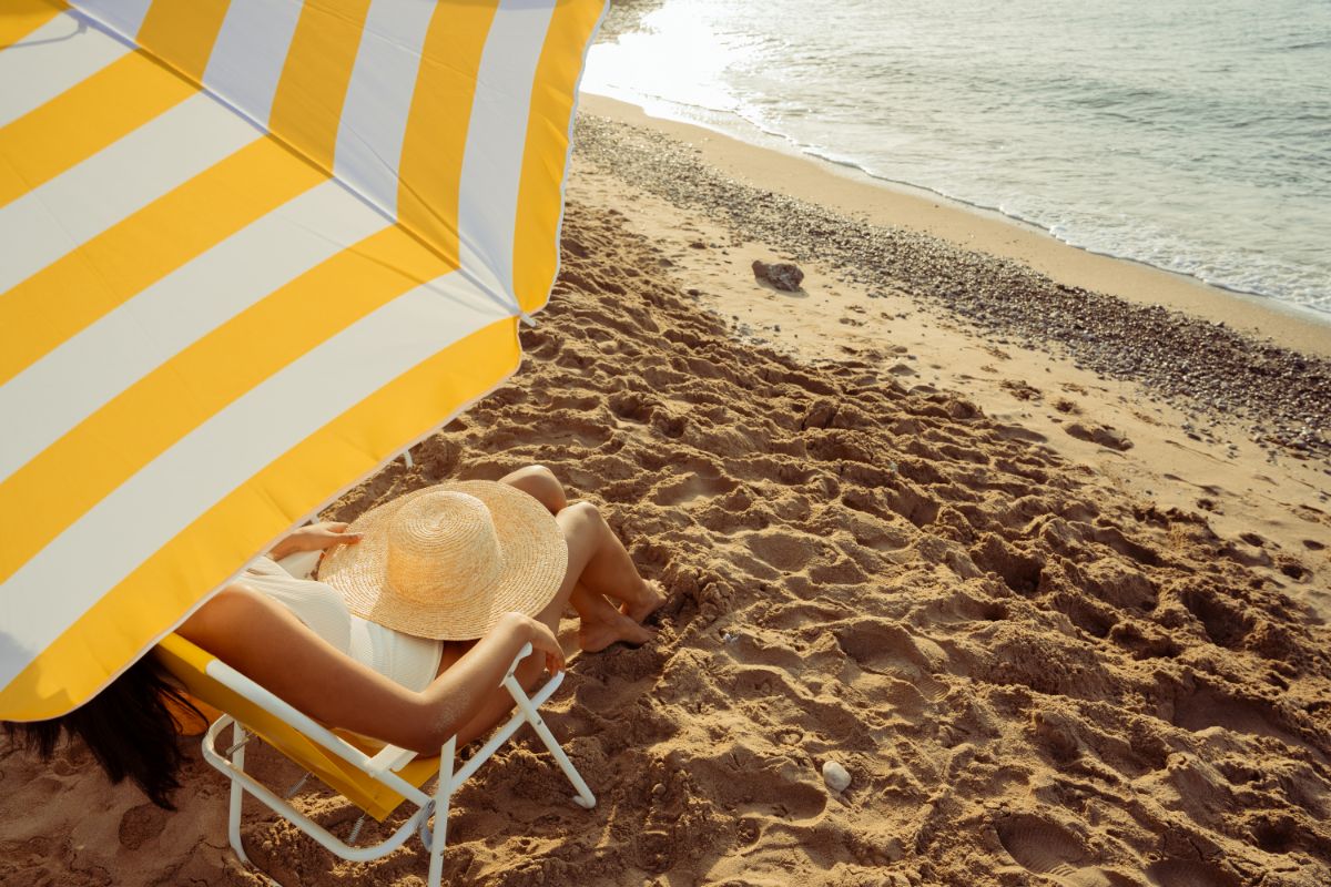 A person enjoys a beach holiday, relaxing on a sandy shore under a yellow and white striped parasol, sitting on a chair with a wide-brimmed hat and gazing at the sparkling sea in the sunlight.