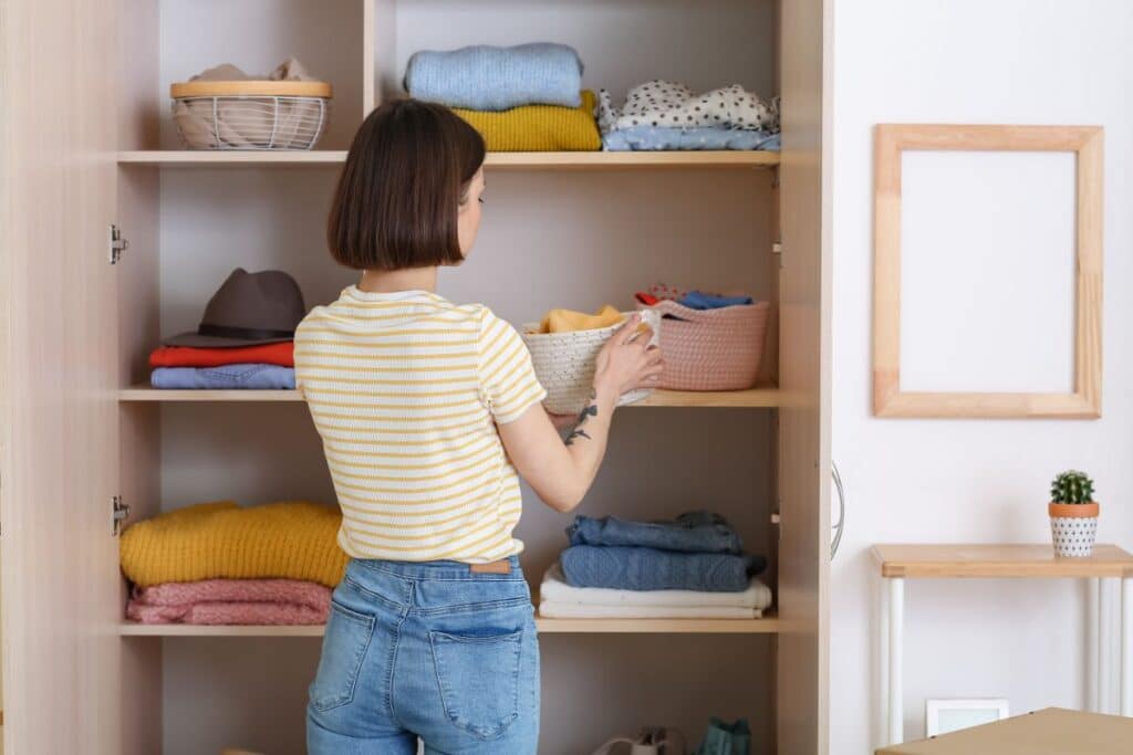 A woman with short brown hair wearing a yellow striped shirt and jeans organises neatly folded clothes on the shelves of a classic wardrobe. She is placing a basket on one of the shelves.