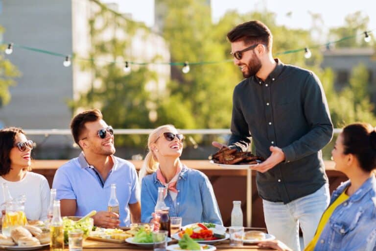 A group of five friends sits at an outdoor table, sharing food and drinks at a lively BBQ party. One man stands holding a plate of grilled meat as the others smile, laugh, and enjoy the sunny weather together.