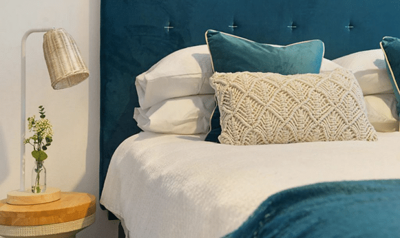 A cosy bed with white linen, textured throw pillows, and a teal headboard sits next to a small wooden bedside table with a glass vase holding flowers and a woven lamp, creating an inviting bedroom sanctuary.