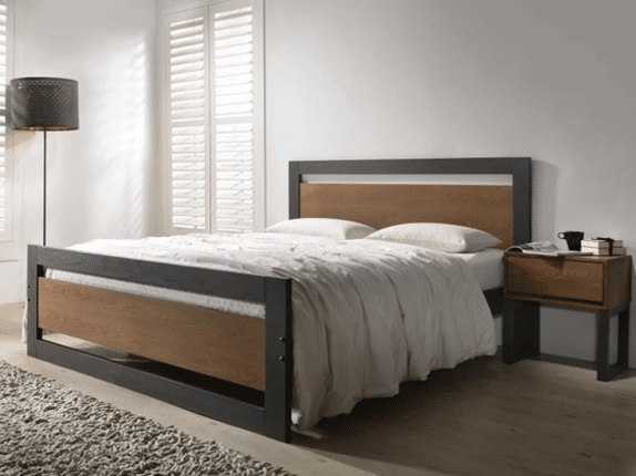 A modern bedroom with a double bed featuring a black metal frame and wooden panels, white bedding, a wooden bedside table with a mug and book by the bed, a floor lamp, and white shuttered windows.
