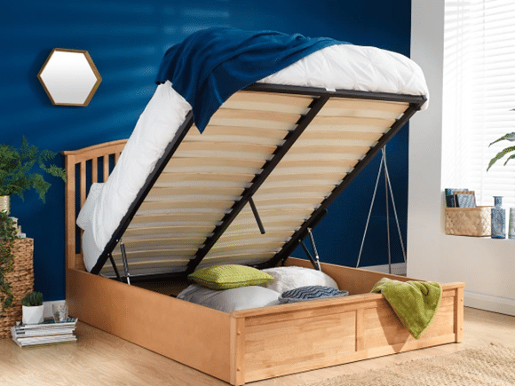 A wooden storage bed with the mattress lifted up reveals ample space underneath for pillows. The room features blue walls, a hexagonal mirror, lush plants, and plenty of natural light streaming in from the window.