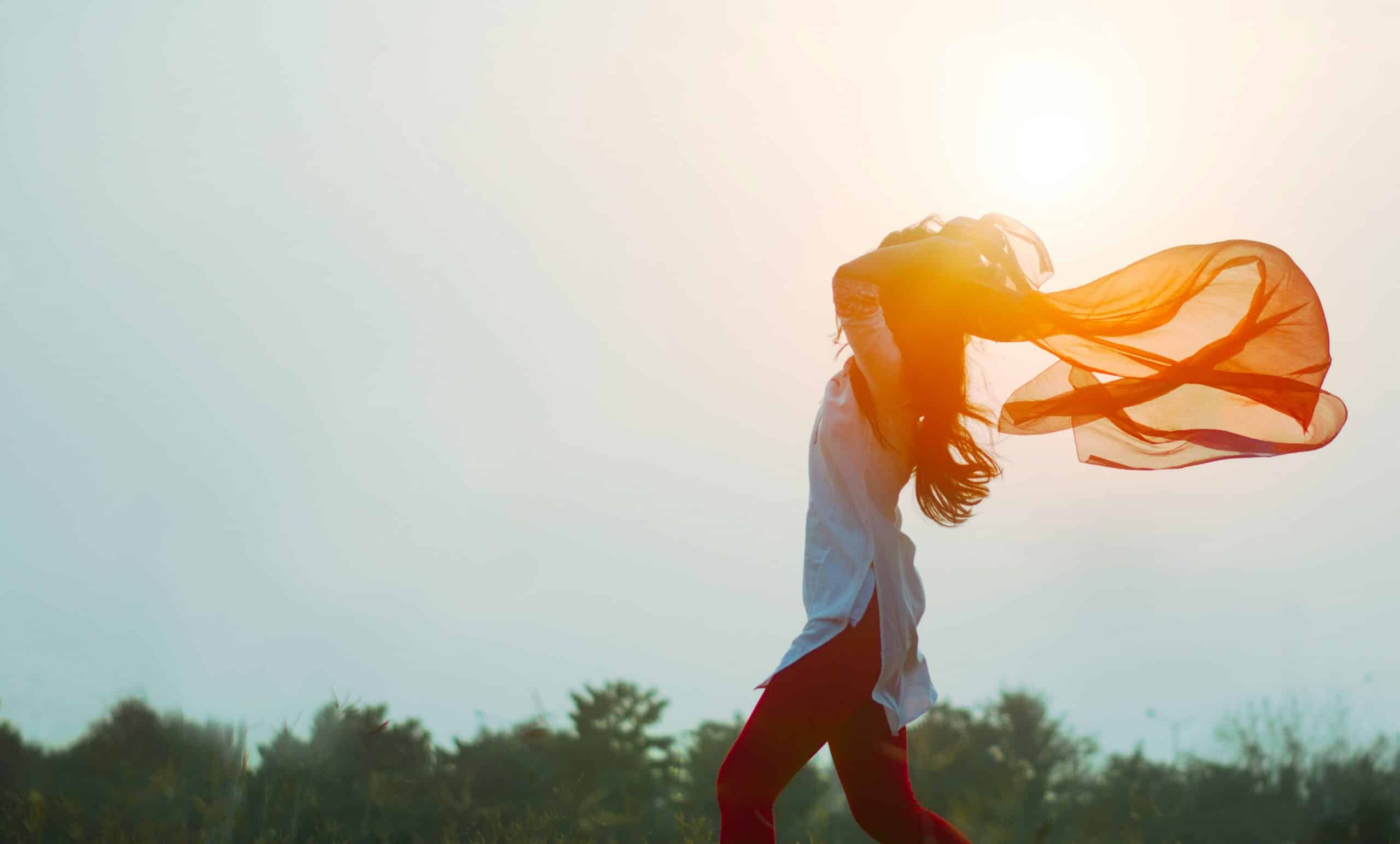 A woman runs outdoors with her arms raised, holding a flowing scarf behind her in the sunlight—capturing the sense of freedom and renewal often felt after divorce. The bright, hazy sky and vibrant plants fill the background.
