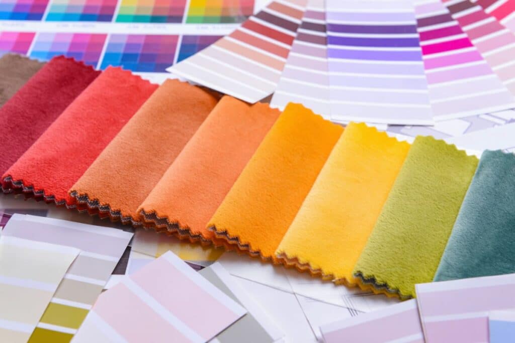 A row of colourful fabric swatches in shades from red to green is displayed on a table, surrounded by various colour sample cards and charts that showcase the latest colour trends and a range of vibrant gradients.